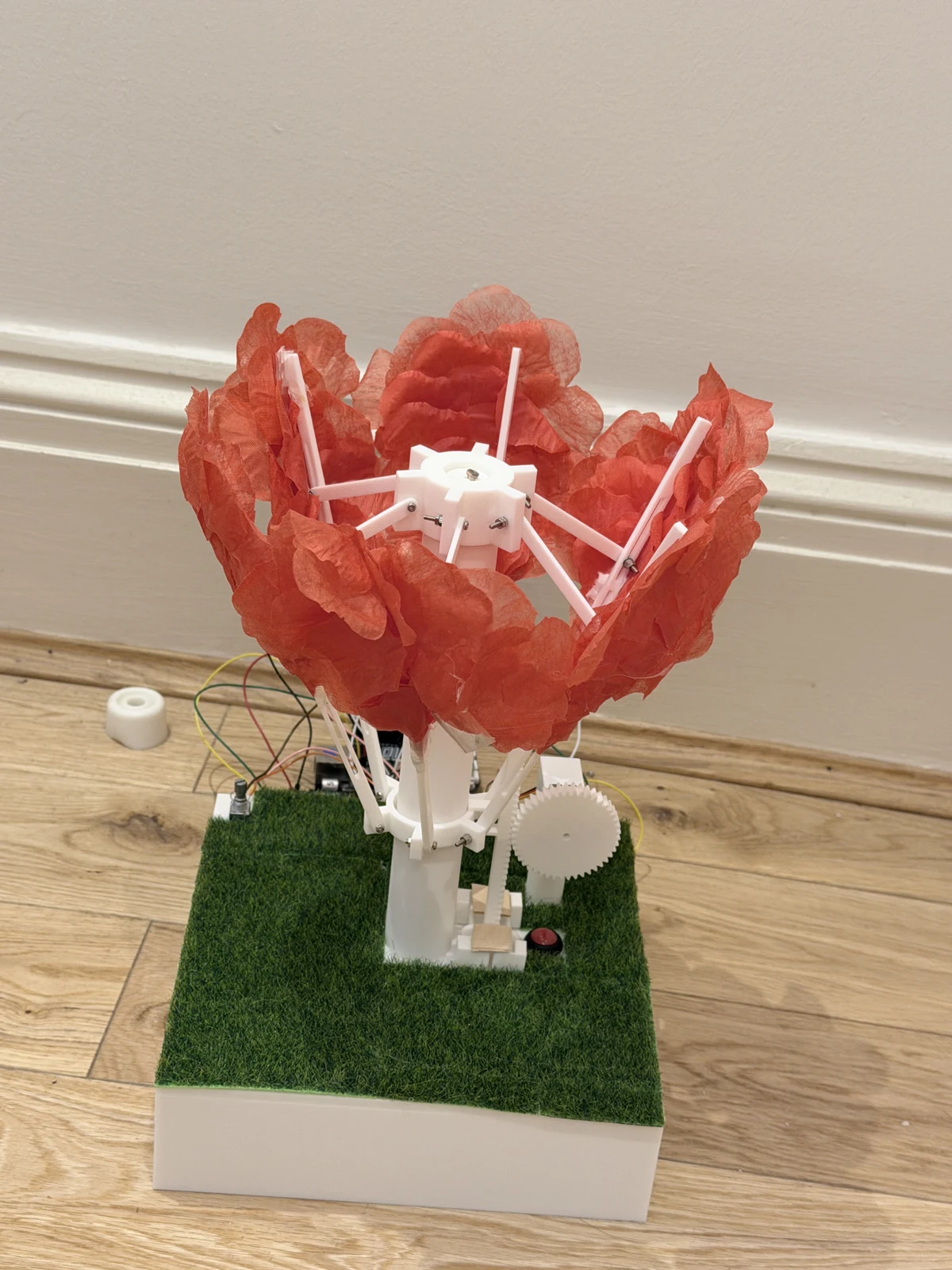Finished ShyBloom prototype in ambient light, with red petals, white printed arms, a central pillar and grass-covered base