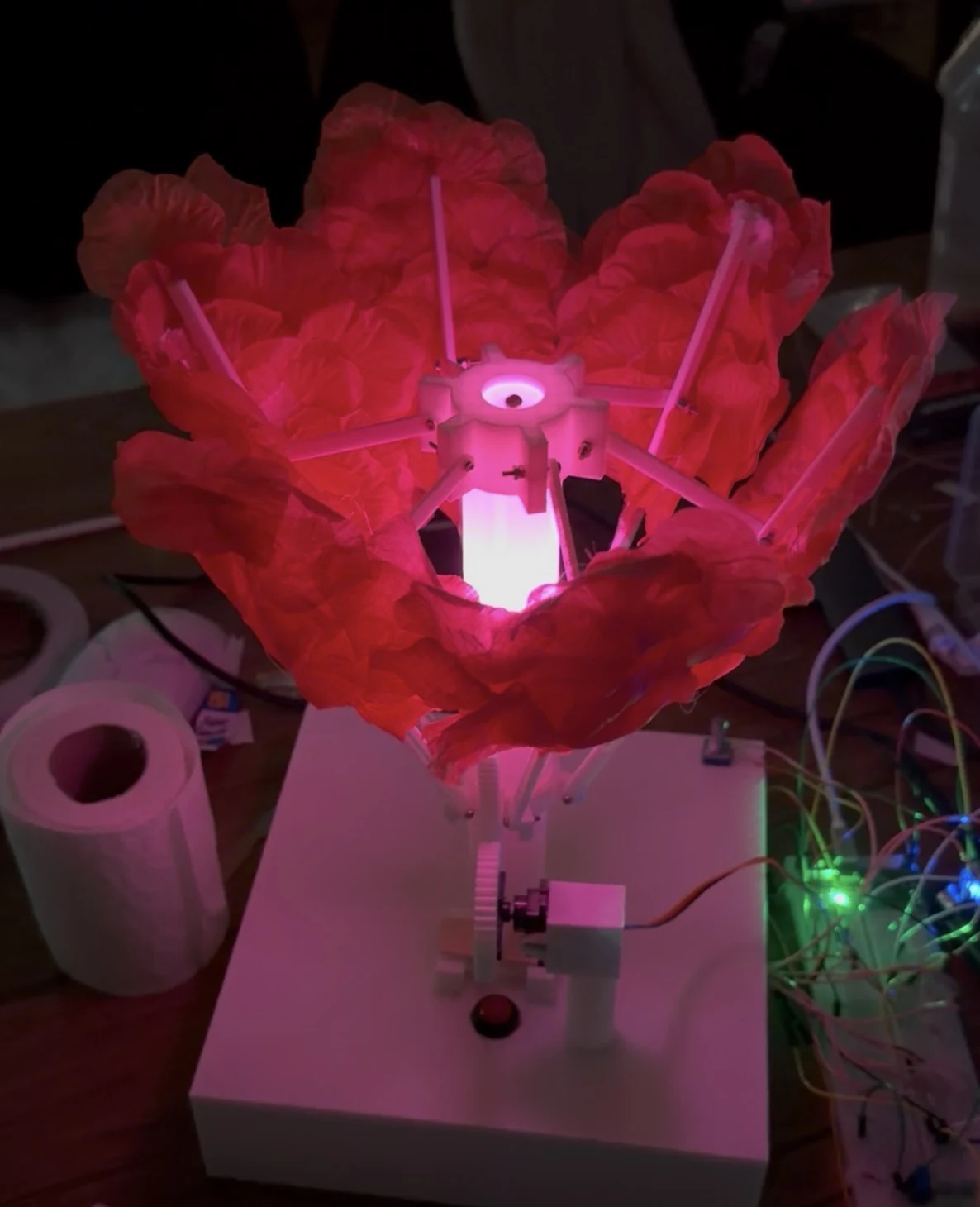 ShyBloom physical prototype glowing pink through red flower petals, with its white linkage and central light column visible